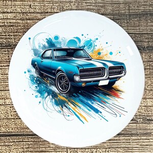 May include: A white ceramic plate featuring a stylized illustration of a classic blue car with white racing stripes. The car is depicted with artistic blue, yellow, and orange accents, creating a dynamic and eye-catching design. The plate has a glossy finish.