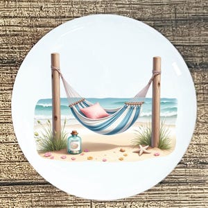 May include: White ceramic plate featuring a beach scene. A blue and white striped hammock hangs between wooden posts on a sandy beach. A pink pillow rests in the hammock, with a turquoise ocean in the background.