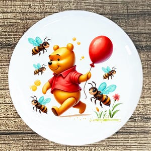 May include: White ceramic plate featuring a cartoon illustration of a yellow bear in a red shirt holding a red balloon, surrounded by bees with blue wings. The plate has a glossy finish and is suitable for serving food.
