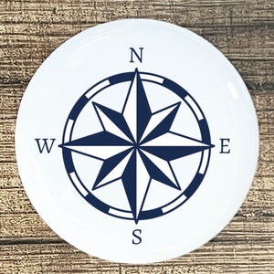 May include: A white round compass with a blue and white star design. The compass points North, South, East, and West.