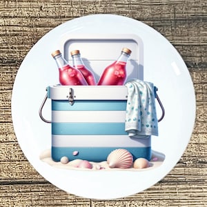 May include: A white ceramic plate features a beach scene illustration. A blue and white striped cooler holds three bottles of pink liquid. A blue and white polka dot towel hangs from the cooler, with seashells and sand in the foreground.