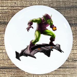 May include: A white ceramic plate featuring a comic book illustration of the Green Goblin. The character is depicted in a green and purple costume, riding a black glider. The plate has a glossy finish.