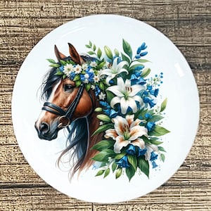 May include: A round white decorative plate featuring a horse's head adorned with a floral arrangement. The horse is brown with a black bridle and mane. The flowers are white lilies, blue bells, and small white and blue blossoms.