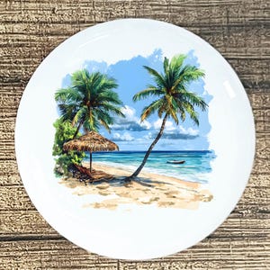 May include: White ceramic plate featuring a tropical beach scene. The image depicts palm trees, a thatched umbrella, lounge chairs, and a small boat on the turquoise water. The sky is blue with fluffy white clouds.