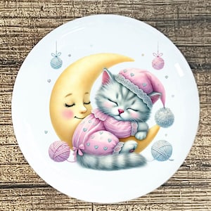 May include: A white ceramic plate featuring a whimsical illustration of a sleeping gray kitten wearing a pink nightcap and pajamas, nestled on a crescent moon. The moon has a smiling face. Pink and blue yarn balls and hanging decorations complete the design.