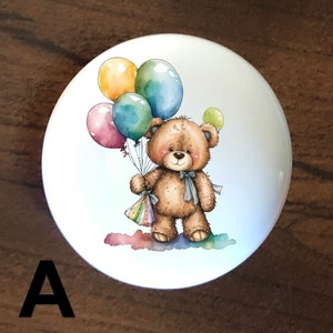 May include: A white ceramic knob with a cartoon illustration of a brown teddy bear holding colorful balloons. The bear is wearing a blue ribbon around its neck.