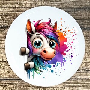 May include: A white ceramic plate featuring a cartoon unicorn with a rainbow mane and big blue eyes. The unicorn peeks from behind a white surface, with colorful paint splatters in the background. The plate has a diameter of approximately 8 inches.