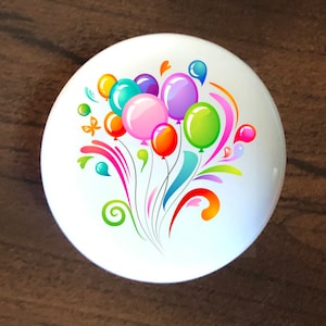 May include: A white ceramic knob with a colorful design of balloons and swirls. The balloons are pink, green, orange, yellow, blue, and purple.
