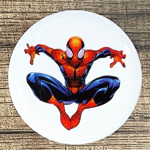 May include: A white round button with a red and blue Spiderman character in a jumping pose. The character is wearing a red and blue suit with a spider logo on the chest.