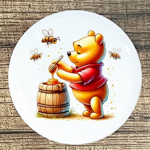 May include: A white ceramic plate featuring a cartoon illustration of Winnie the Pooh in a red shirt, scooping honey from a wooden barrel. Three bees are flying around the scene. The plate has a glossy finish.