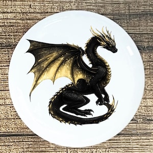 May include: A white, round decorative plate featuring a black dragon with gold accents. The dragon has large, detailed wings and a long tail. The plate is set against a wooden background.