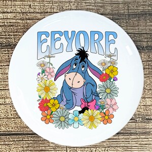 May include: A white round button with a blue and purple cartoon donkey surrounded by colorful flowers. The text "EEYORE" is written in a blue and purple gradient at the top of the button.