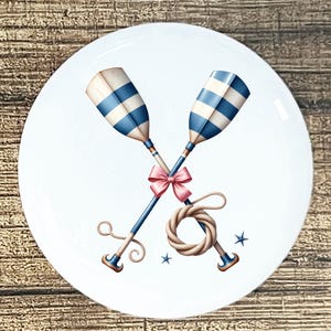 May include: White ceramic plate featuring crossed oars with blue and white stripes, tied with a pink bow and rope. The design includes small blue star accents, evoking a nautical theme. The plate has a glossy finish.