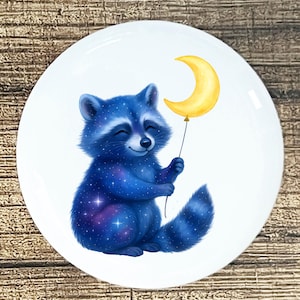 May include: A white ceramic plate featuring a smiling raccoon with a galaxy-themed fur pattern. The raccoon holds a yellow crescent moon balloon. The design is whimsical and colorful, perfect for a child's room or kitchen.