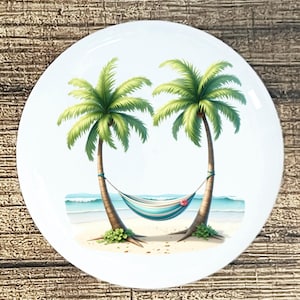 May include: White ceramic plate featuring a tropical beach scene. Two palm trees frame a striped hammock, with a sandy beach, ocean, and sky in the background. The palm trees have green fronds and brown trunks.