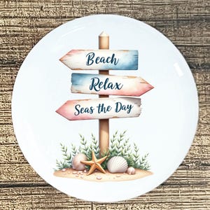 May include: White ceramic plate with a beach-themed design. The plate features a wooden signpost with three directional arrows that read "Beach", "Relax", and "Seas the Day". The design includes seashells, a starfish, and greenery.