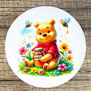 May include: White ceramic plate featuring a cartoon illustration of Winnie the Pooh in a red shirt, holding a honey jar. Colorful flowers and bees surround the bear. The jar has the word "HONEY" on it.