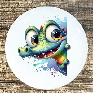 May include: A white ceramic plate featuring a cartoon alligator with large, expressive eyes and a friendly smile. The alligator is primarily green and blue, with red accents and a rainbow-colored belly. Watercolor-style splashes surround the character.