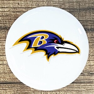 May include: A white, round coaster featuring the Baltimore Ravens logo. The logo depicts a stylized raven's head in purple, black, and gold, with a red eye and the letter "B" in the center. The coaster is likely made of ceramic or a similar material.