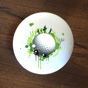 May include: White ceramic knob with a green, black, and white golf-themed design. The design features a golf ball, golf players, and green splatters.