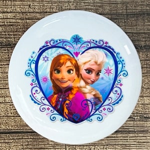 May include: White ceramic plate with a blue and purple heart-shaped design featuring the characters Anna and Elsa from the movie Frozen. The design includes snowflakes and swirls.