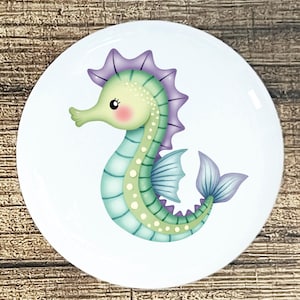 May include: A white, round ceramic plate featuring a cartoon seahorse illustration. The seahorse is depicted in shades of green, blue, and purple, with a pink blush on its cheek. The plate is set against a wooden background.