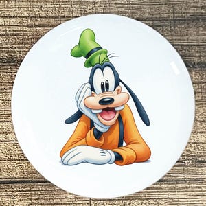 May include: White ceramic plate featuring a cartoon image of Goofy. Goofy is depicted with a green hat, orange shirt, black suspenders, and white gloves. The plate has a glossy finish and is suitable for serving food.