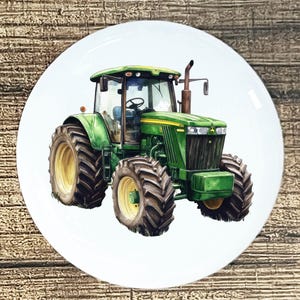 May include: A white ceramic plate featuring a detailed illustration of a green tractor with large tires. The tractor has yellow accents and is set against a white background. The plate is round and appears to be a decorative item.
