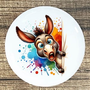 May include: White ceramic plate featuring a cartoon donkey with wide blue eyes and an open mouth, peeking from behind a white surface. Colorful watercolor paint splatters in shades of blue, orange, and purple decorate the plate.