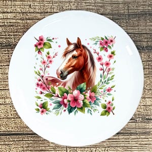 May include: White ceramic plate featuring a detailed illustration of a brown and white horse's head surrounded by pink cherry blossoms and green leaves. The horse has a flowing mane and is set against a square floral frame.