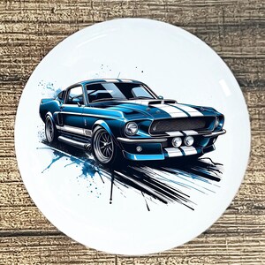 May include: A blue classic car with white racing stripes is depicted on a white circular plate. The car has a detailed illustration with blue paint splatter accents. The plate is set against a wooden background.