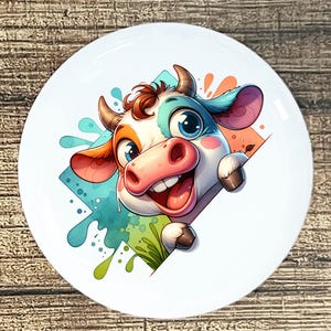 May include: A white ceramic plate featuring a cartoon cow with a cheerful expression. The cow has a colorful design with blue, orange, and pink accents. The plate has a glossy finish and is set against a wooden background.