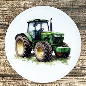 May include: A white ceramic plate featuring a watercolor painting of a green John Deere tractor. The tractor has large black tires with yellow rims and is set against a backdrop of green grass. The plate is round.