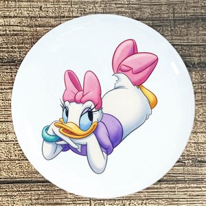 May include: A white, round decorative plate featuring a cartoon illustration of Daisy Duck. Daisy is depicted in a relaxed pose, wearing a pink bow, purple top, and turquoise bracelet. The plate has a glossy finish.