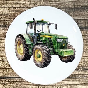 May include: A white ceramic plate featuring a watercolor illustration of a green tractor with yellow wheels. The tractor has a detailed design, including headlights, a cab, and a smokestack. The plate is set against a wooden background.