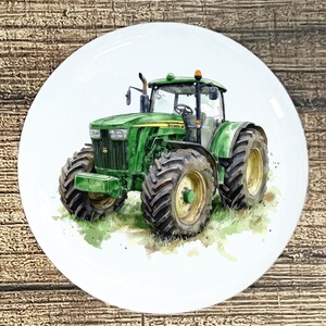 May include: A white ceramic plate featuring a watercolor illustration of a green tractor. The tractor has large black tires with yellow rims and is set against a backdrop of green grass. The plate is round and has a glossy finish.