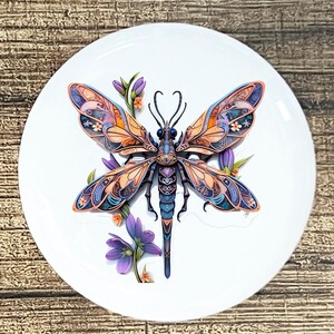 May include: A white ceramic plate featuring a detailed dragonfly design. The dragonfly has orange, blue, and purple wings, with a matching body. Purple flowers and green leaves surround the dragonfly, creating a nature-inspired theme.