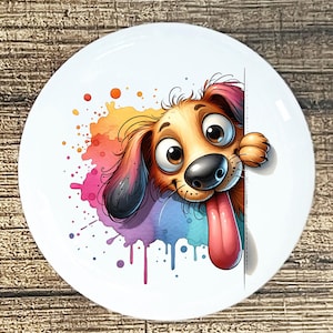 May include: White ceramic plate featuring a cartoon dog with brown fur, big eyes, and a long pink tongue. The dog is peeking from behind a white surface, with colorful watercolor paint splatters in the background.