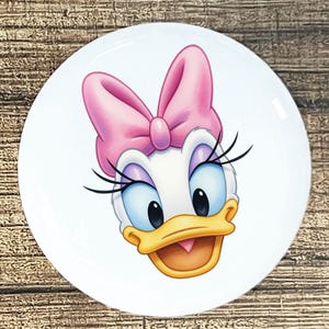 May include: A white ceramic plate featuring a cartoon image of Daisy Duck. Daisy has a pink bow, blue eyes, long eyelashes, and a yellow beak. The plate is round and sits on a wooden surface.