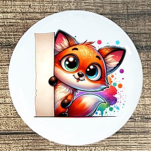 May include: A white ceramic plate featuring a cartoon fox with orange fur, big blue and green eyes, and a fluffy tail. The fox is peeking from behind a blank sign, with colorful paint splatters in the background.