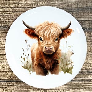 May include: A white ceramic plate featuring a detailed illustration of a Highland cow. The cow has long, shaggy, light brown fur, dark horns, and is set against a backdrop of soft green and brown foliage.