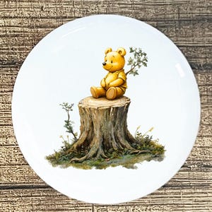 May include: A white ceramic plate featuring a yellow bear sitting on a tree stump. The bear is wearing a yellow shirt and is surrounded by greenery. The plate has a glossy finish and is suitable for serving food.