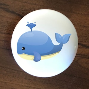 May include: A white ceramic knob with a blue whale illustration. The whale is cartoon-like and has a small spout on its head. The whale is facing right and has a light blue body with a white belly.