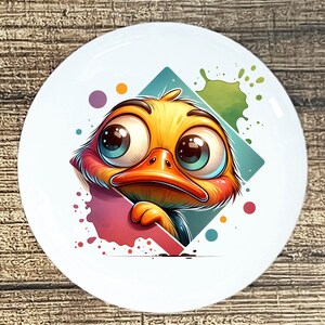 May include: White ceramic plate featuring a cartoon duck with large, expressive eyes. The duck is yellow, orange, and red, set against a teal diamond and colorful paint splatters. A whimsical design for kitchenware or home decor.