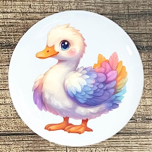 May include: A white ceramic plate featuring a cartoon duckling with a white body, orange beak and feet, and large blue eyes. The duckling has colorful feathers in shades of purple, blue, pink, and orange. The plate is round and has a glossy finish.