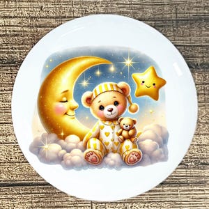 May include: A white ceramic plate featuring a whimsical illustration of a teddy bear in pajamas, sitting on a cloud with a smaller teddy bear, a smiling crescent moon, and a star. The design has a soft color palette of yellow, blue, and white.