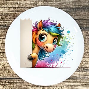 May include: A white ceramic plate featuring a cartoon unicorn with a rainbow mane and blue eyes. The unicorn peeks from behind a blank white space, with colorful paint splatters in the background. The plate is on a wooden surface.
