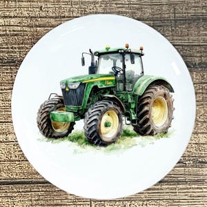 May include: A white ceramic plate featuring a watercolor painting of a green tractor with large tires. The tractor is set against a backdrop of green grass. The plate has a glossy finish and a round shape.