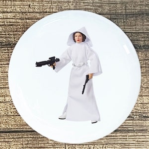 May include: A white plastic action figure of Princess Leia from Star Wars. She is wearing a white hooded robe and holding a blaster in one hand and a pistol in the other.