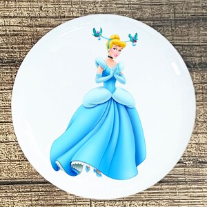 May include: A blue and white Cinderella button featuring a cartoon image of Cinderella in her iconic blue ball gown with a tiara and two blue birds on her head.